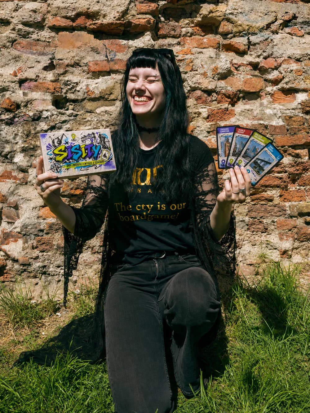 Prowadząca UrbGames z pudełkiem gry SISITY i talią kart przy ceglanym murze
